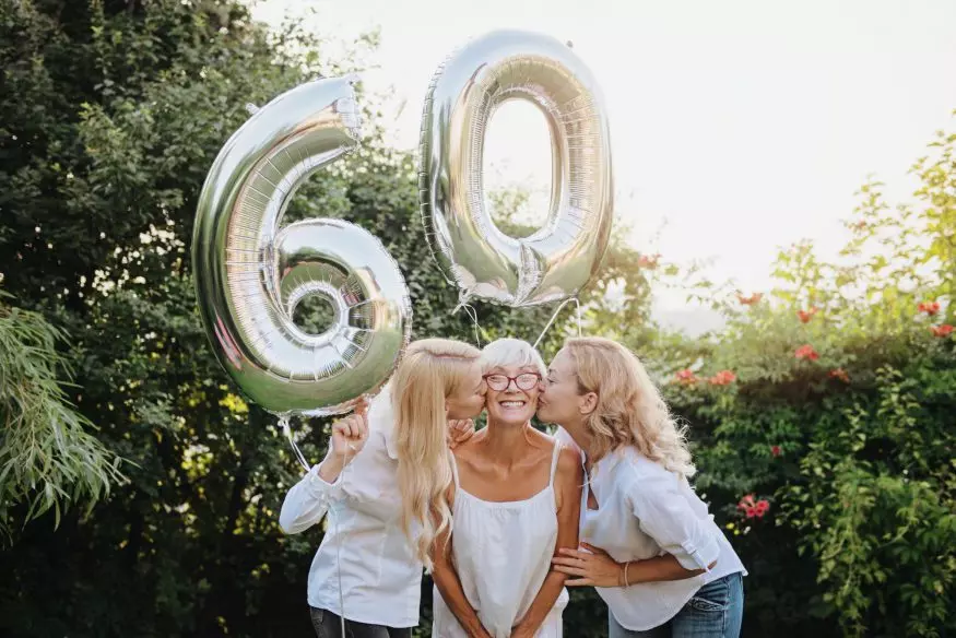 60th birthday party ideas: Daughters kissing their mother on the cheek while holding number 60 balloons