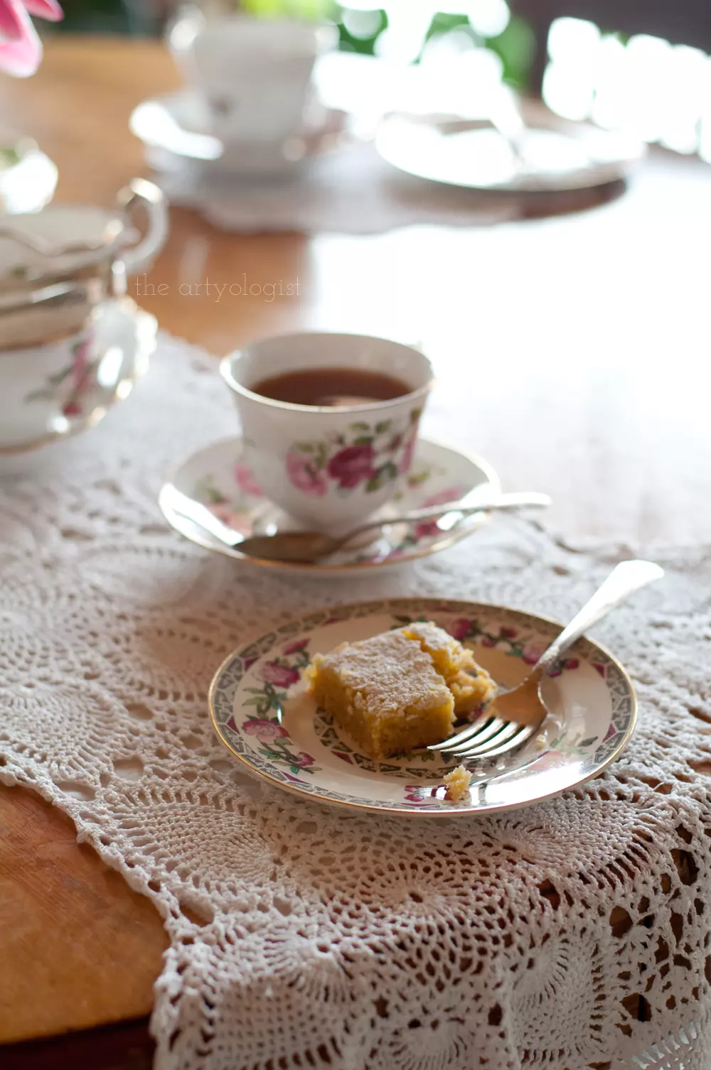 A small plate with a lemon square on it and a cup of tea in the background