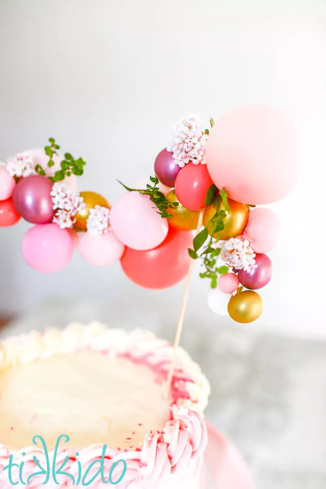 Miniature balloon garland cake topper made with pink and gold balloons and small flowers, on top of a pink cake.