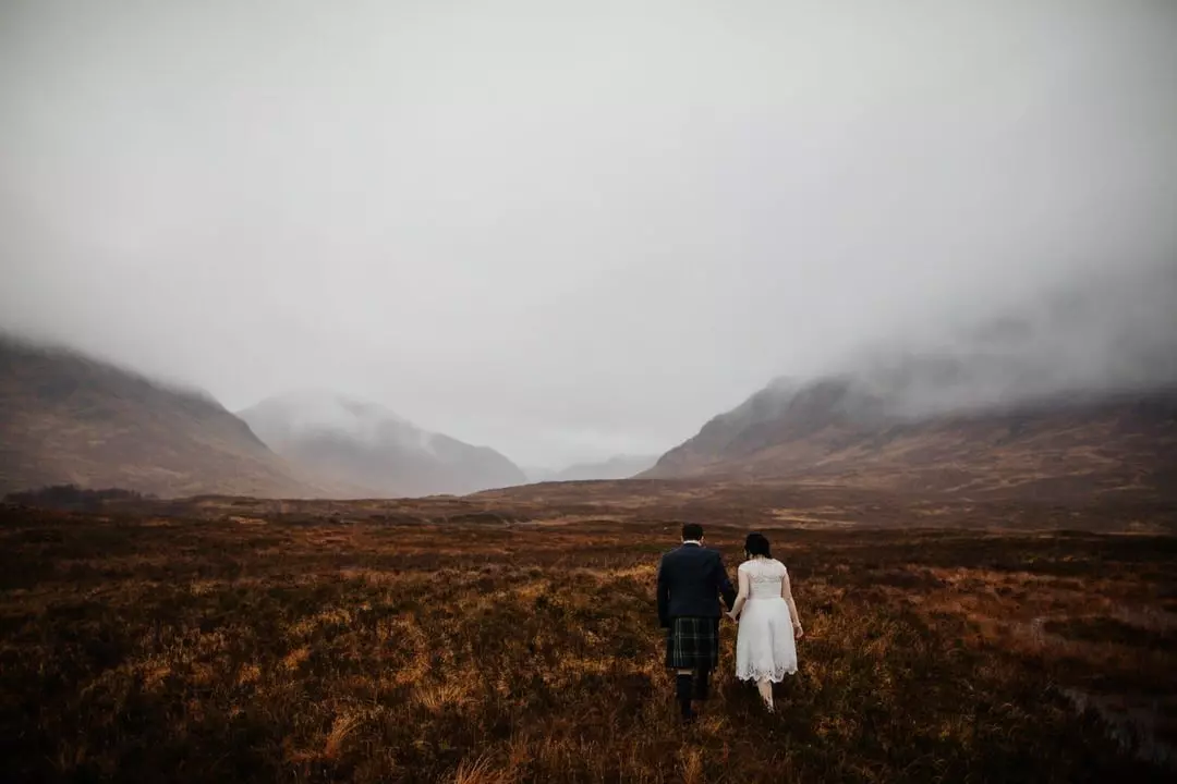 Bride and groom walking across a misty field with the hills of Glencoe in the background