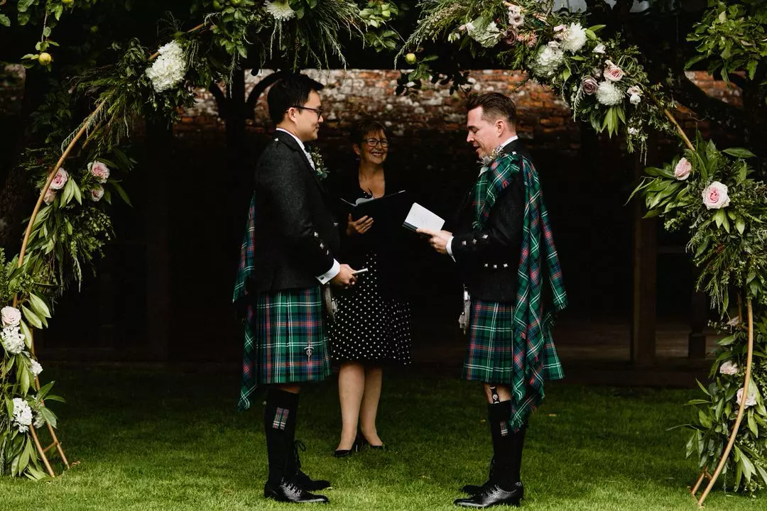 Male same-sex couple reading vows to each other at their wedding ceremony under floral arch